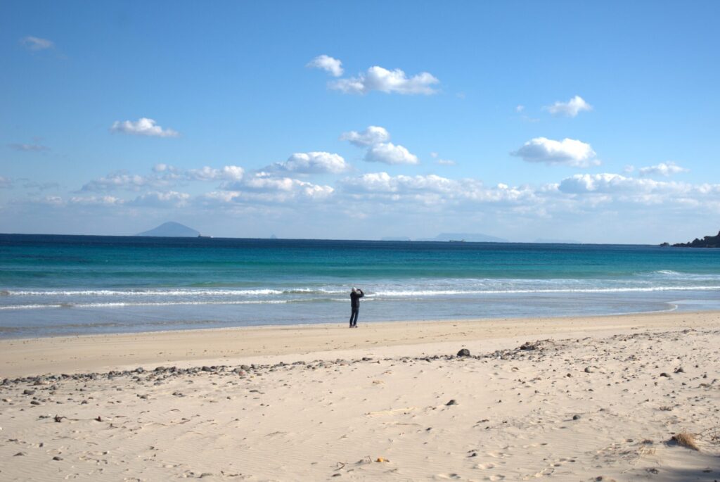 青空の下、白浜大浜海水浴場の広い砂浜が広がっています。遠くには青い海と、水平線に浮かぶ島々が見えます。海岸の手前には、小石が点在する砂浜が続き、1人の人物が海に向かって立っている様子が写っています。穏やかな波が浜辺に打ち寄せ、静かな雰囲気が漂っています。