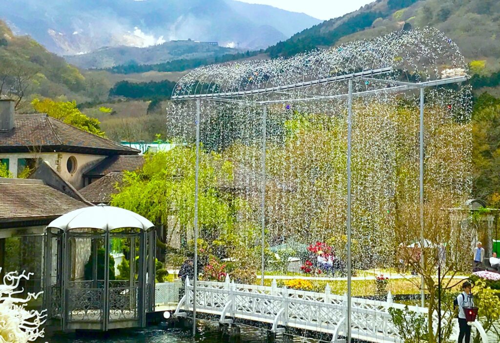 箱根ガラスの森美術館の風景。山に囲まれた美しい谷間に位置し、クリスタル・ガラスのアーチの光の回廊が輝く庭園が見えます。白い装飾が施された小さなパビリオンや、池にかかる白い橋があり、その周囲には緑豊かな木々や色とりどりの花が咲いています。背景には霞がかかった山々が広がっています。