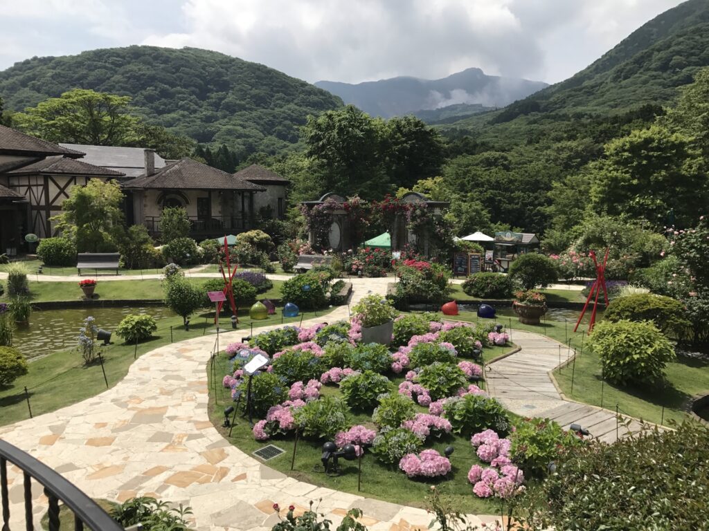 箱根ガラスの森美術館の庭園。緑豊かな山々に囲まれた風景が広がり、手入れの行き届いた庭園には色とりどりの花が咲いています。中央にはピンク色のアジサイが植えられ、石畳と木製の小道が交差しています。左側には小さな池があり、右側には赤い彫刻が立っています。背景には木造の建物やアーチ型の門が見え、山の間には雲がかかっています。