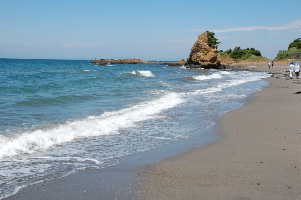 神奈川県横須賀市の立石公園の砂浜と海岸線。青い波が穏やかに打ち寄せ、遠くに立石の岩がそびえ立つ風景。晴天の下、散策を楽しむ人々の姿も見られる。