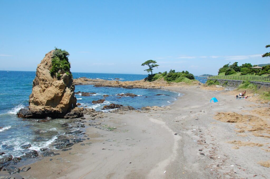 神奈川県横須賀市の立石公園の風景。青い海と広がる砂浜、特徴的な立石の岩がそびえ立つ絶景スポット。遠くには緑の丘と松の木が見える。