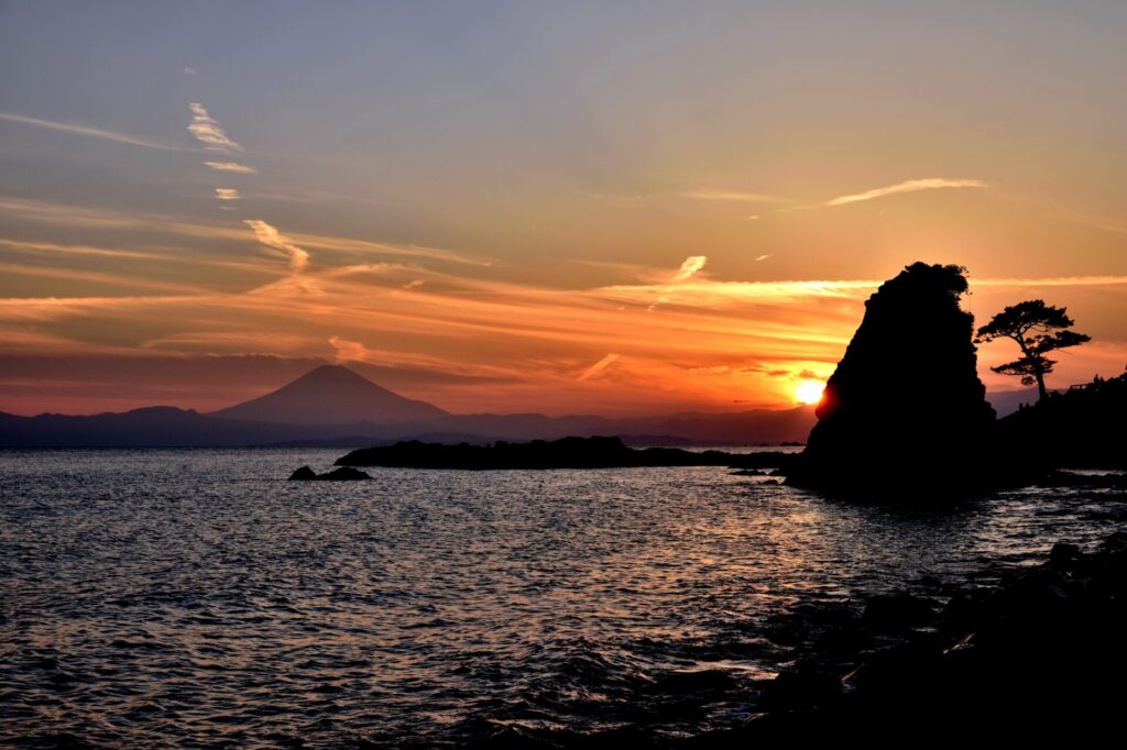 神奈川県横須賀市・立石公園の夕景。富士山のシルエットと赤く染まる空、海に沈む太陽が織りなす幻想的な風景。手前には波打つ海、右側に立石のシルエットが際立つ。