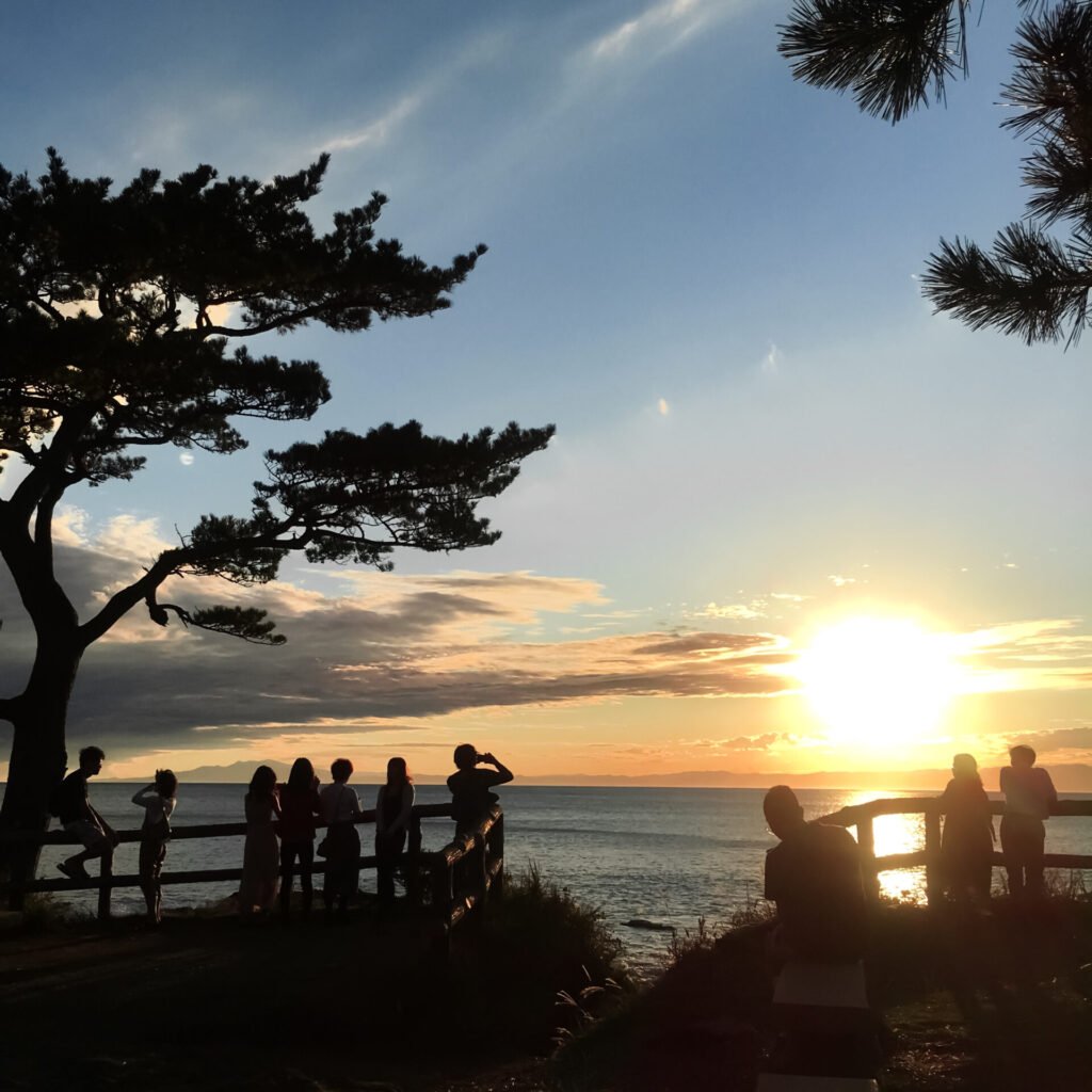 神奈川県横須賀市・立石公園の夕景。沈む夕日を背景に、シルエットになった人々と松の木が美しく映える。ロマンチックな雰囲気の絶景スポット。