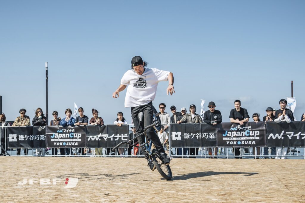 Sasaki, a BMX rider, performing at the Mynavi Japan Cup BMX Freestyle event. Spectators watch as he demonstrates technical flatland skills at the 2023 competition in Yokosuka City.