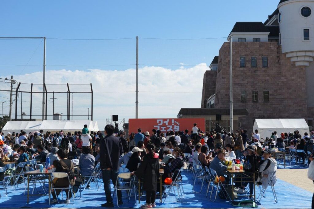 Photo of Y! Festa Oppama showing a large crowd gathered at the outdoor venue with seating and tables, enjoying the event under clear skies. A stage is visible in the background with the event banner.